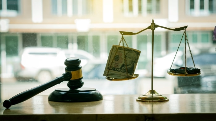 Scales of justice with money and a car lying inside and a lawyer hammer on the table